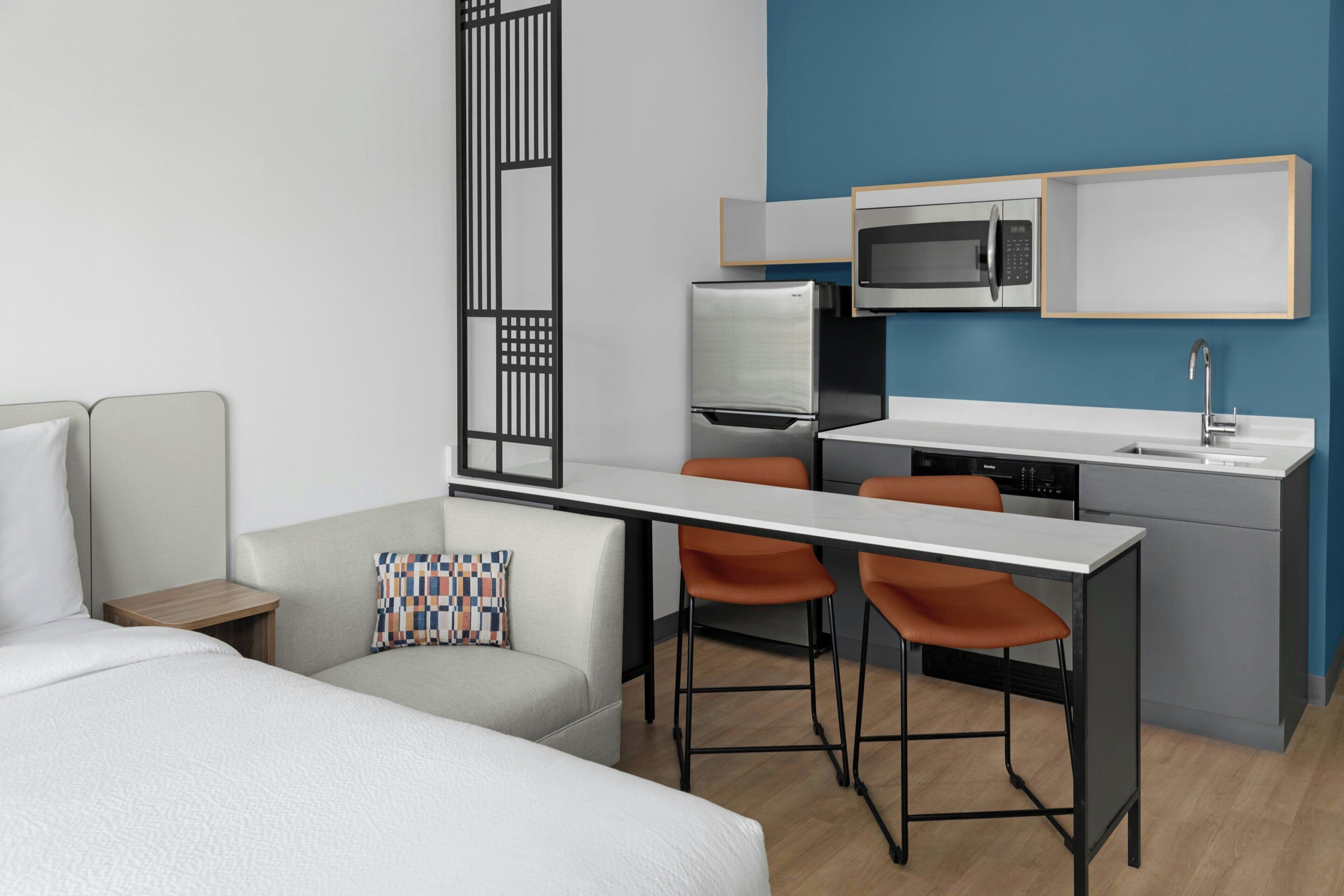 A modern hotel room with a bed, a sofa, a small table, a kitchenette with a microwave, a refrigerator, and two bar stools.