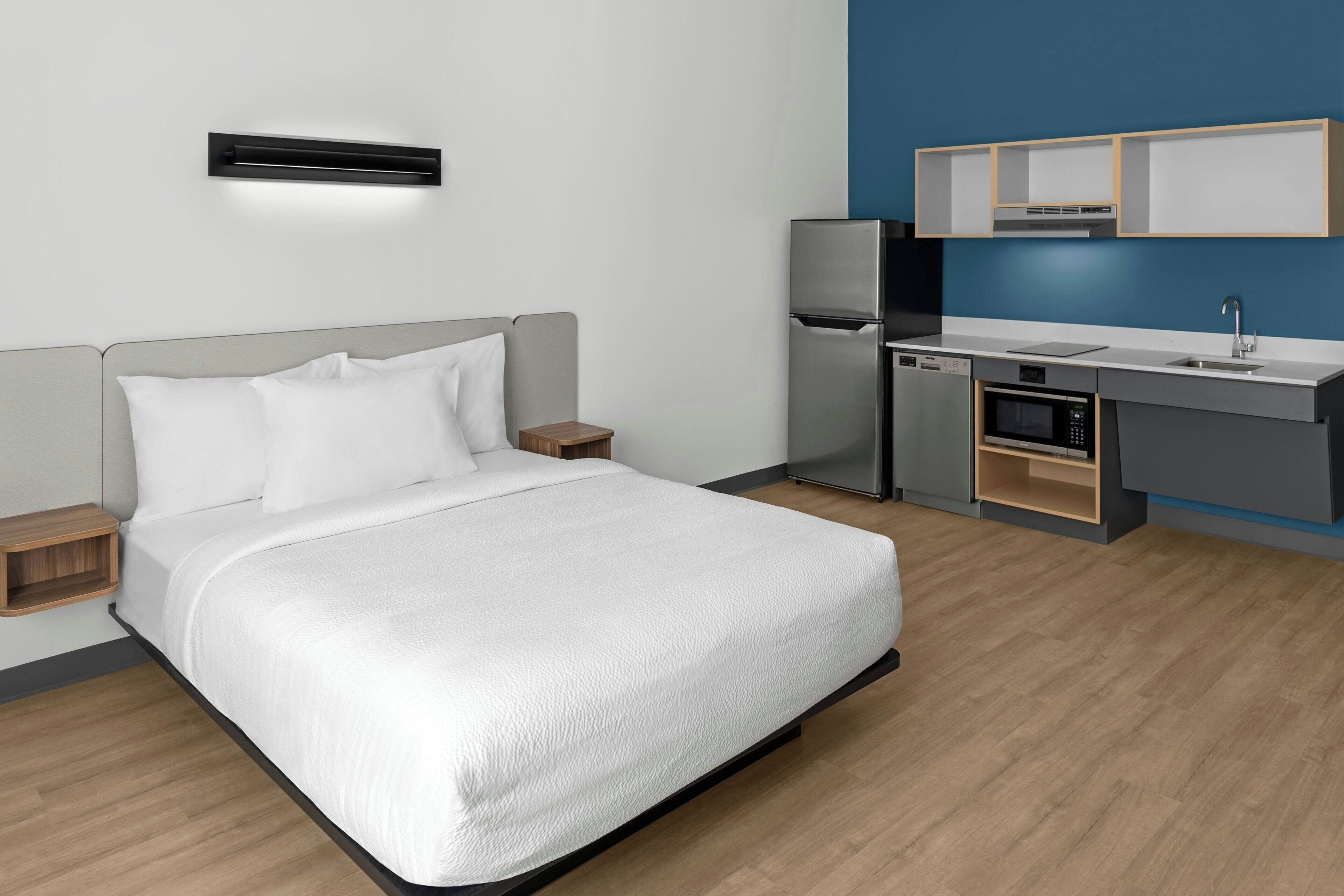 A modern hotel room with a bed, bedside tables, a built-in air conditioning unit, a compact kitchenette with a refrigerator, microwave, and sink.