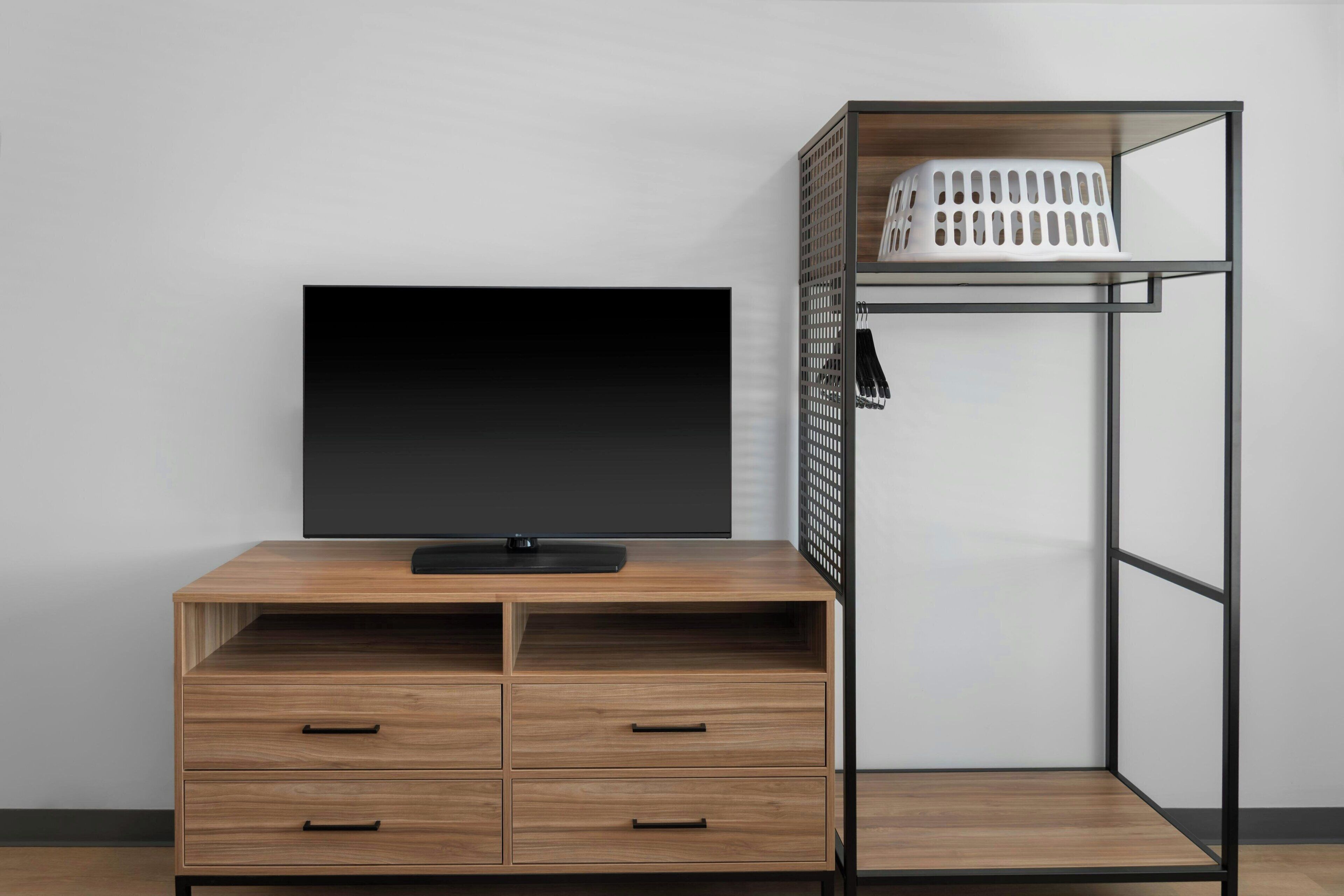 A room with a wooden desk, a flat-screen TV, and a metal shelving unit with a laundry basket.