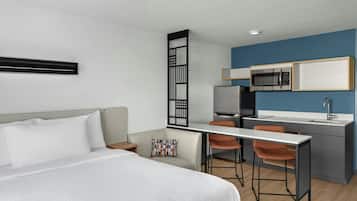 Una habitación de hotel moderna con cama, escritorio, dos sillas, kitchenette y un panel decorativo negro.