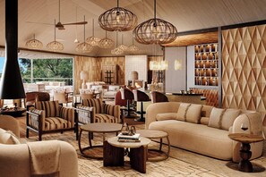 A spacious living area with a large sofa, striped armchairs, and a coffee table. There are wicker pendant lights and a bookshelf filled with books and decorative items. - Kinara, A Luxury Collection Safari Camp, Masai Mara (Maasai Mara)