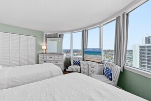 A bedroom with a large bed, a dresser, a chair, and a view of the beach. - Peck Plaza 9NW (Daytona Beach Shores)
