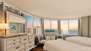 Ein Hotelzimmer mit einem großen Bett, einem Schminktisch, einem Schrank und Blick auf den Sonnenuntergang über dem Meer.