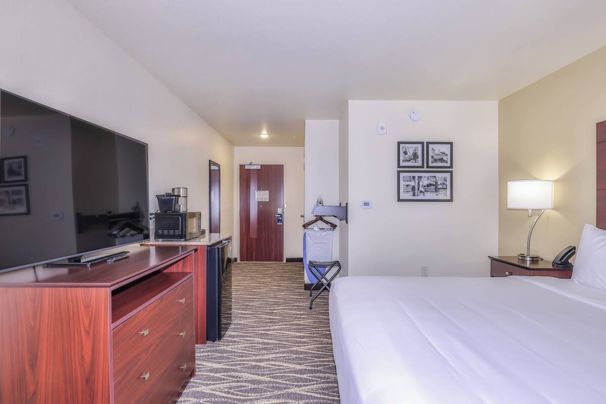 Habitación de hotel con cama, escritorio con televisor de pantalla plana, microondas, cafetera, lámpara y armario.