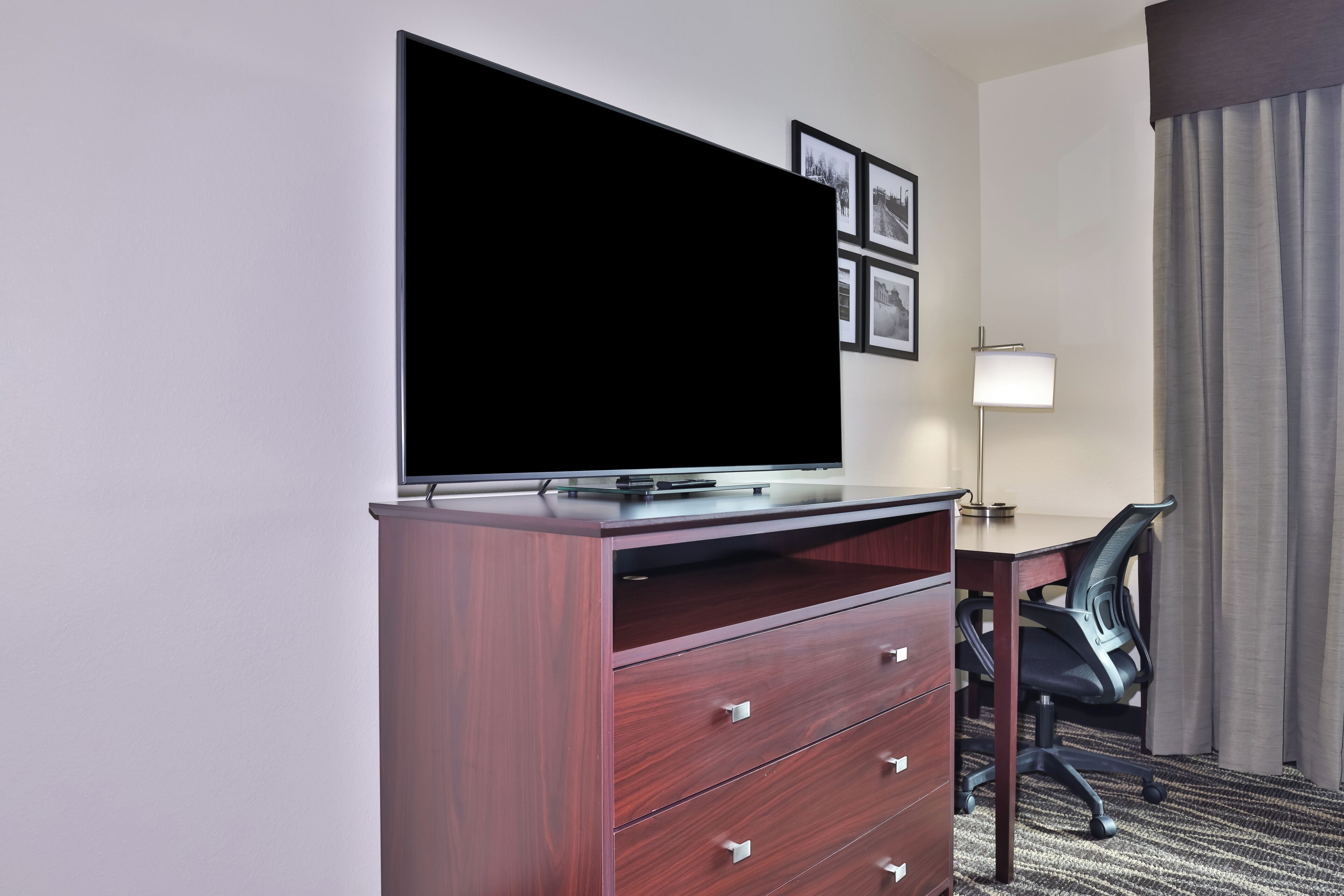 A hotel room with a flat-screen TV on a wooden cabinet, a desk with a chair, and a lamp.