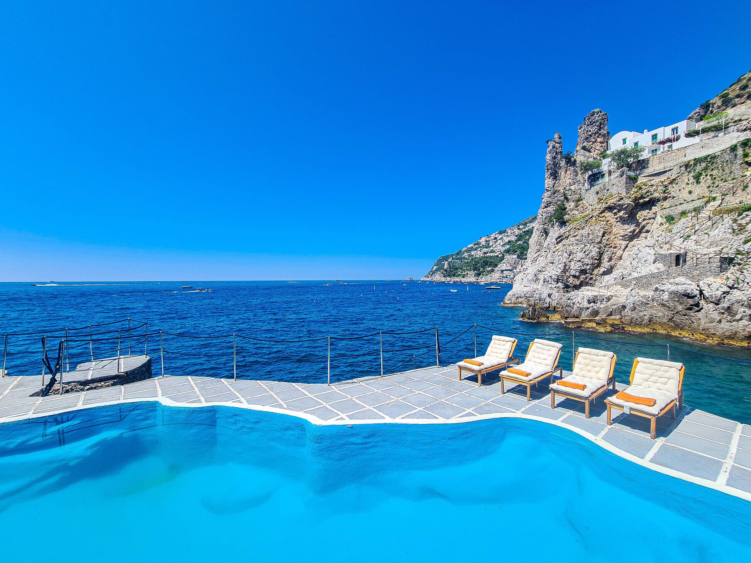 Ein Poolbereich mit Liegestühlen und Blick auf das Meer und schroffe Klippen.