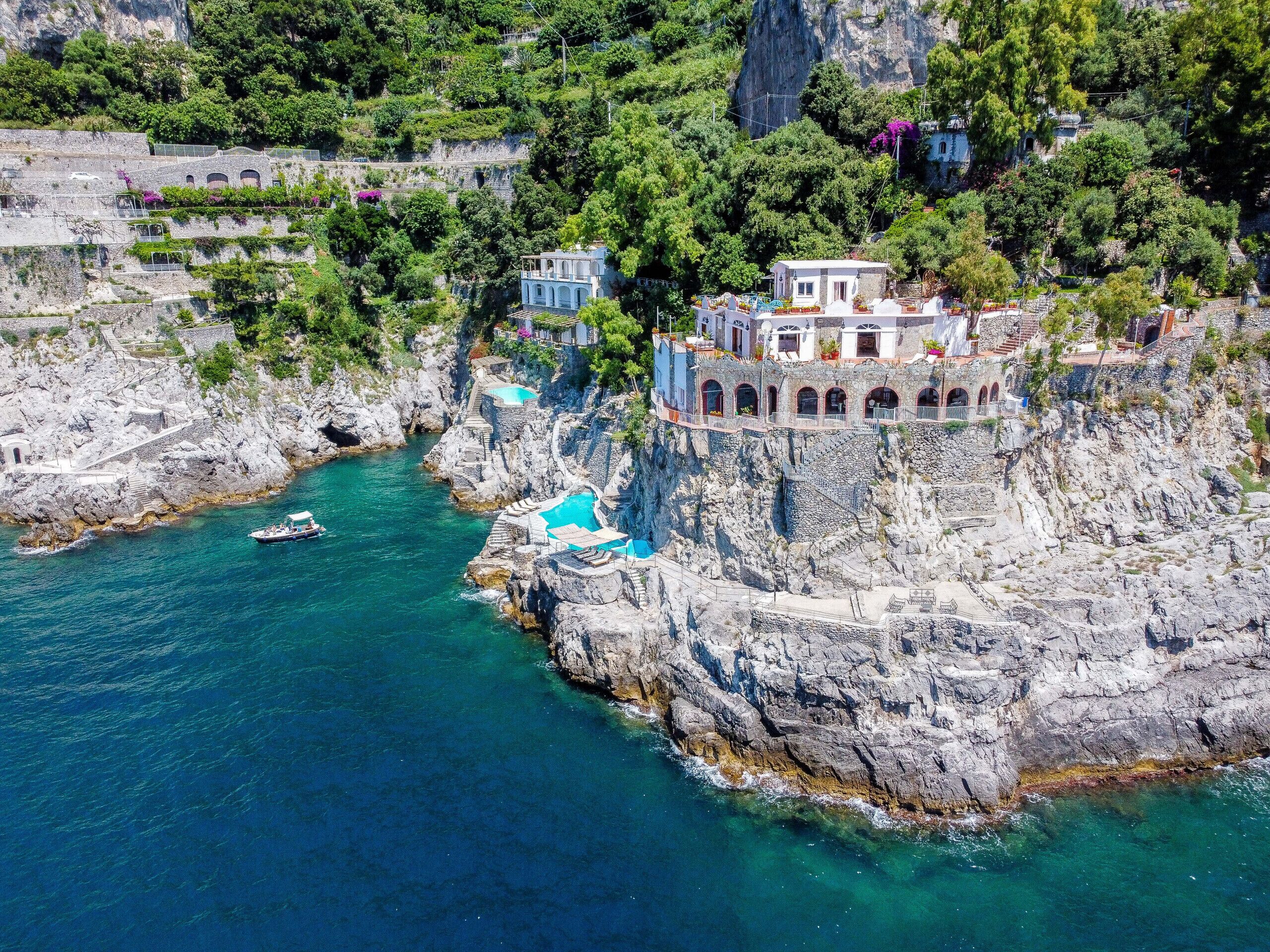 A coastal cliffside hotel with multiple buildings, a swimming pool, and a boat in the water.