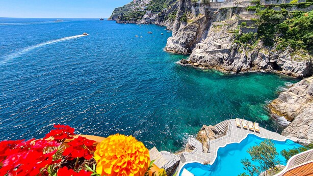 Blick auf die Küste mit blauem Meer, einem Felsen und einem Schwimmbecken mit weißer Brüstung.