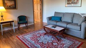 Ein Wohnzimmer mit grauem Sofa, einem Holztisch, einem gemusterten Teppich, einem Holzstuhl, einer Lampe und einer Tür zu einem anderen Raum.