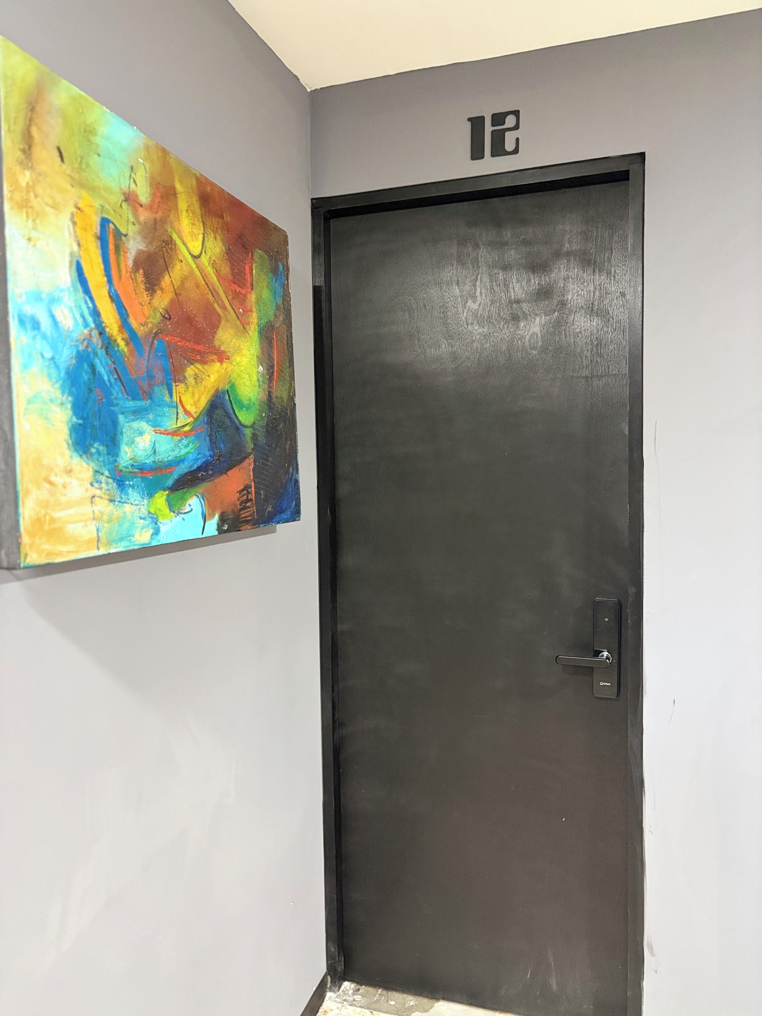 Una puerta con una placa de número, una puerta negra y un cuadro abstracto de colores en la pared.