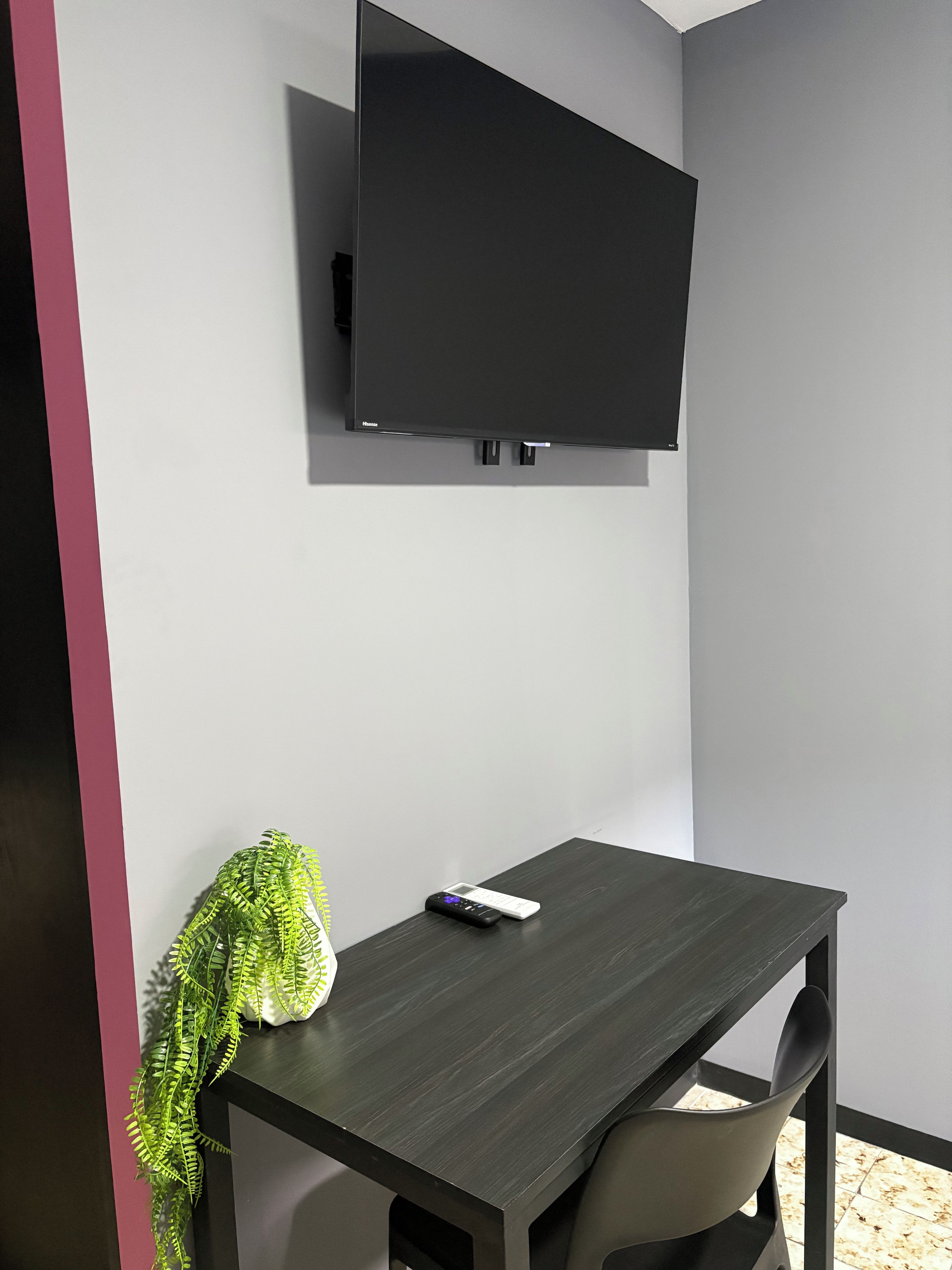 Una habitación con televisión de pantalla plana montada en la pared, una mesa de madera oscura, una silla y una planta en maceta.