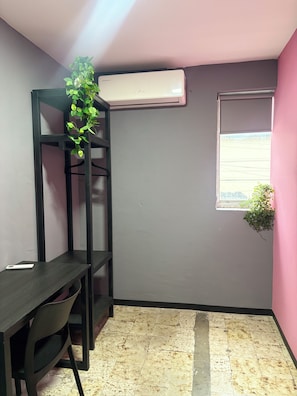A room with a desk, a black shelving unit, an air conditioner, a window, and a plant.