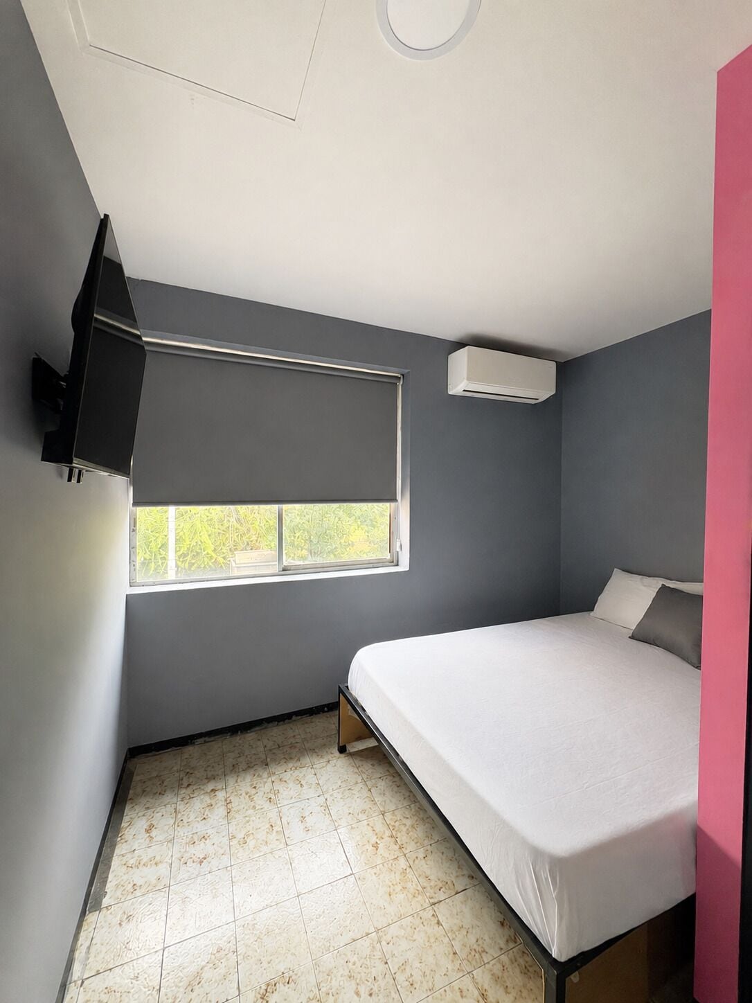 A bedroom with a bed, a window, a wall-mounted air conditioner, and a partial view of a pink wall.