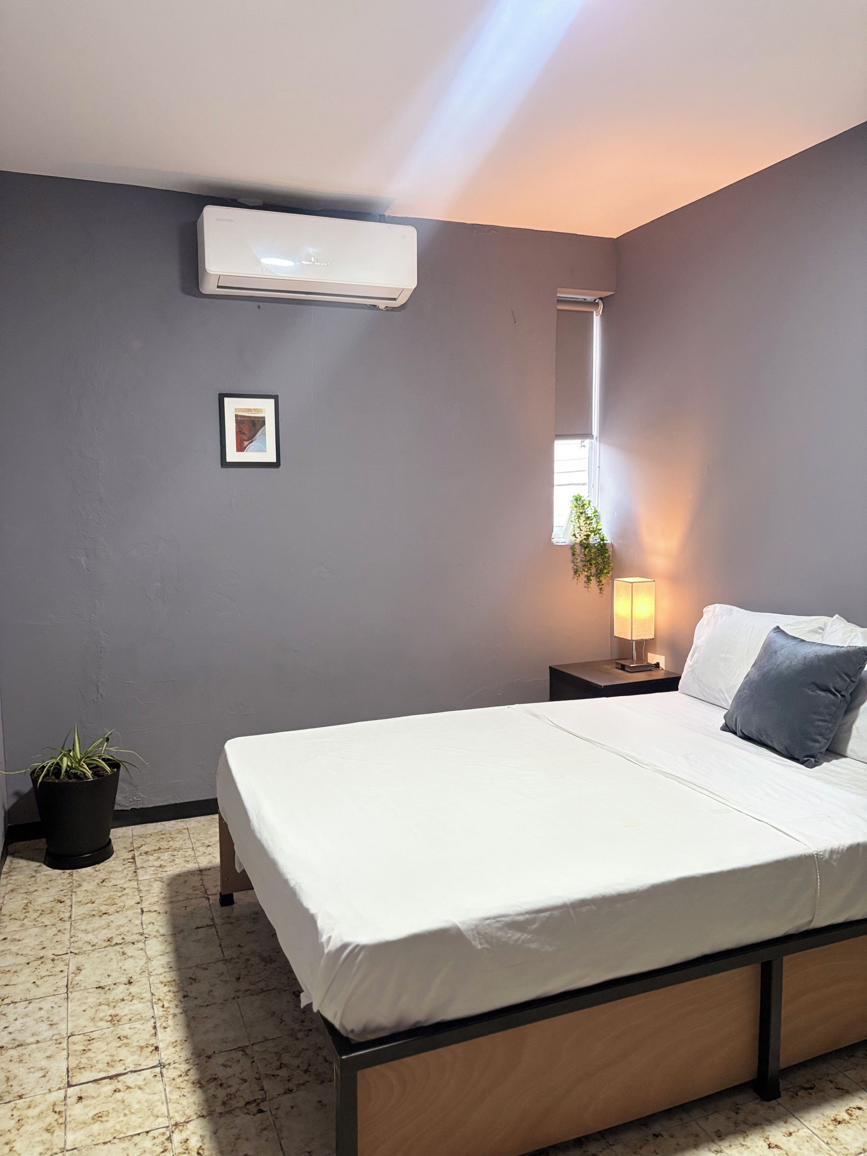 A bedroom with a bed, a nightstand, a lamp, a potted plant, and an air conditioning unit.