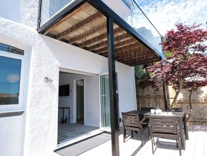 Ein modernes Haus mit überdachter Terrasse, einer Glasschiebetür, die zu einem Innenraum führt, und einem kleinen Tisch mit Stühlen im Freien.