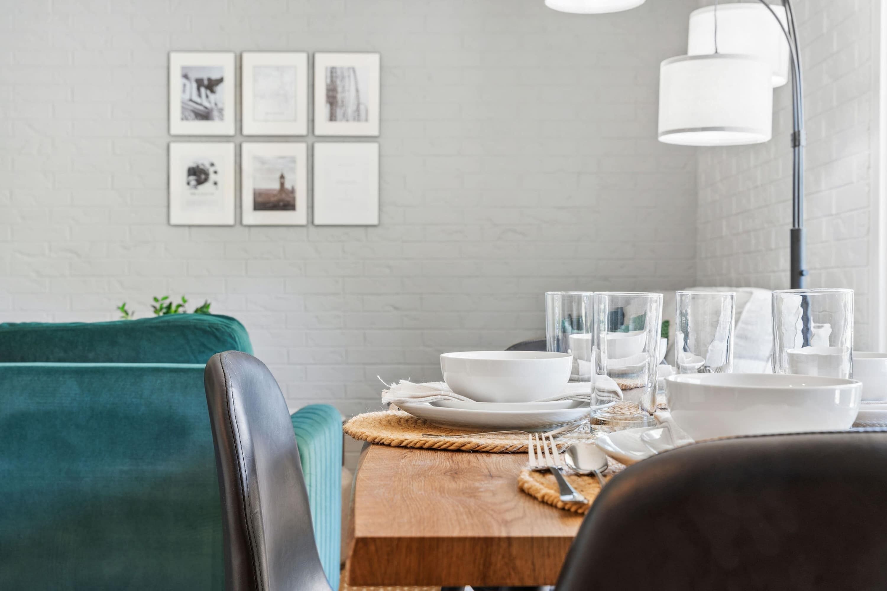 Un comedor con una mesa de madera dispuesta para cuatro, platos, cuencos y vasos blancos. Se observan cuadros en la pared y un sofá verde al fondo.