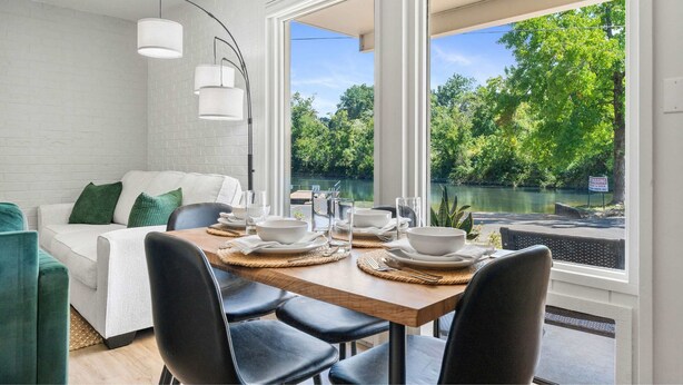 A dining area with a table set for four, large windows offering a view of a lake and greenery, and a white brick wall. - Cozy Creekside Landing (Branson)