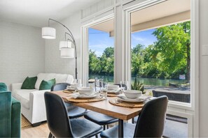 Un coin repas avec une table dressée pour quatre personnes, de grandes fenêtres donnant sur un lac et la verdure environnante, et un mur en briques blanches.