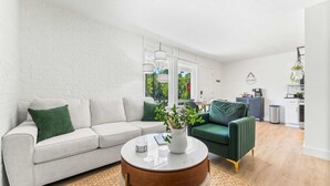 A modern living room with a white brick wall, a grey sofa, a green armchair, a round coffee table, and a kitchen area in the background. - Cozy Creekside Landing (Branson)