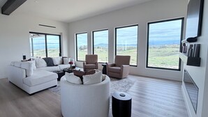 Een ruime woonkamer met een grote witte bank, twee fauteuils en een rond poef. Vier grote ramen bieden uitzicht op een wijngaard.