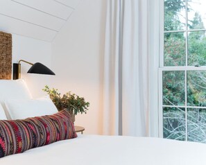 A bedroom with a bed, a bedside lamp, a window with curtains, and a view of trees. - Kubli Haus (Jacksonville)