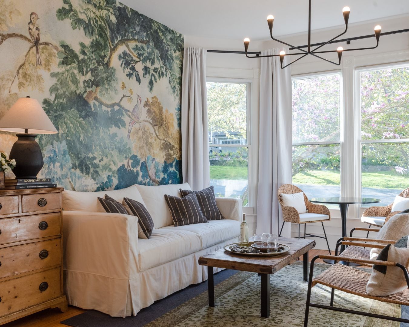 A room with a white sofa, a wooden coffee table, wicker chairs, a large window with curtains, and a wall mural.