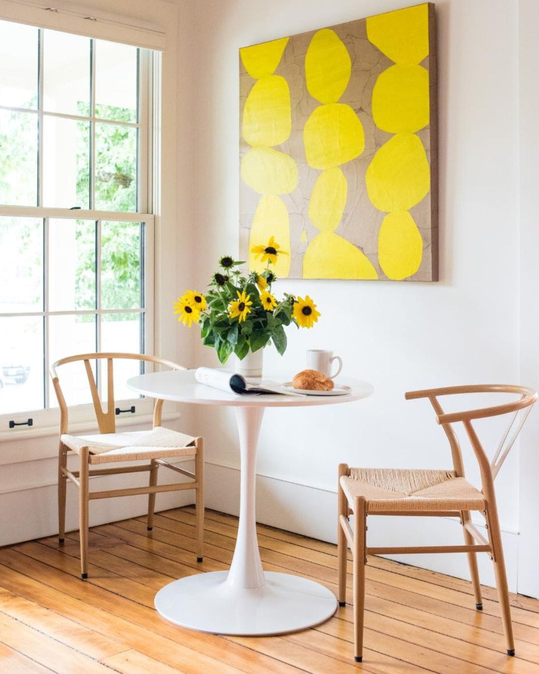 A room with a round white table, two wooden chairs, a vase with sunflowers, and a yellow abstract painting on the wall.
