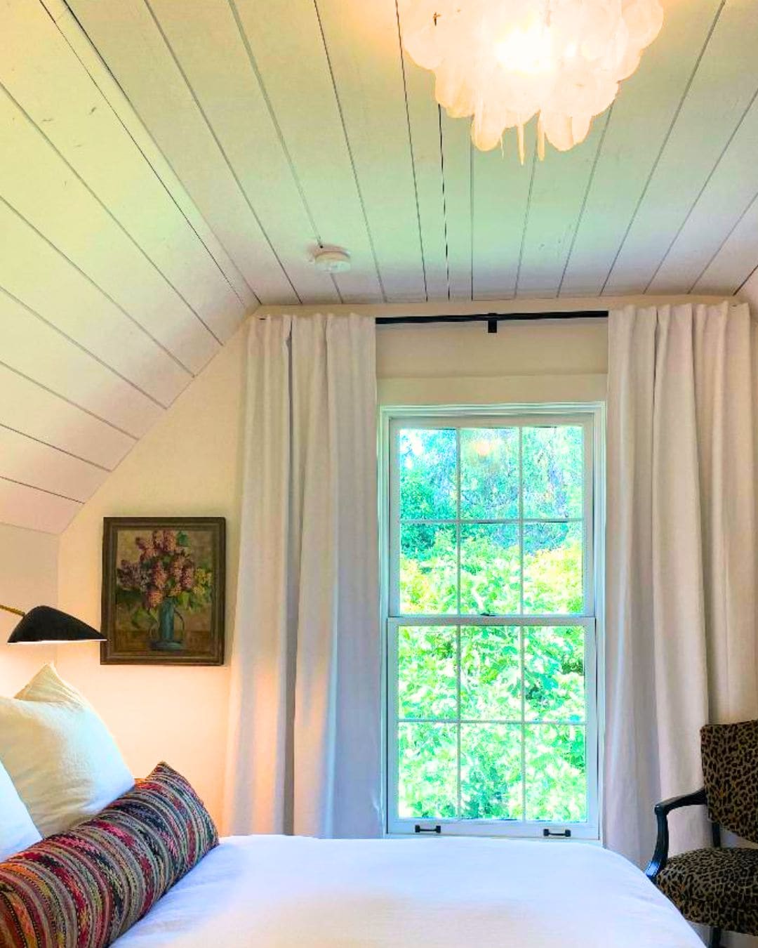 Un dormitorio con cama, una silla, un ventanal con cortinas, un cuadro y una lámpara en el techo.