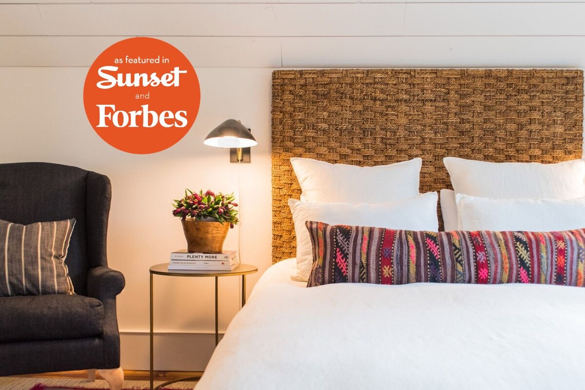 A hotel room with a bed, a chair, a side table with a lamp, and a wall-mounted light fixture.