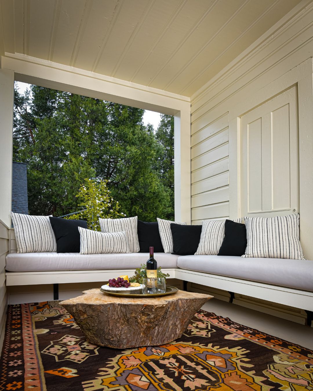 A covered outdoor seating area with a sofa, a table with a bottle and snacks, and a patterned rug.