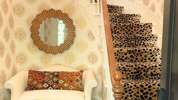 Una escalera con alfombra de estampado de leopardo, un sofá blanco con cojines estampados, un espejo redondo y una araña.