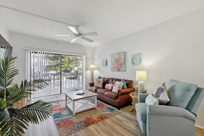 A spacious living room with a brown leather sofa, a grey armchair, a coffee table, and a ceiling fan. There are two wall-mounted lamps and a large window with blinds. - Beach Escape With Direct Beach Access (Ormond Beach)