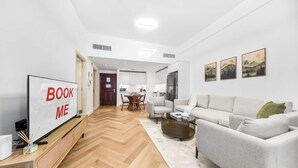 Een moderne woonkamer met een bank, een salontafel en een tv op een standaard. Achtergrond toont een eethoek met een tafel en stoelen.