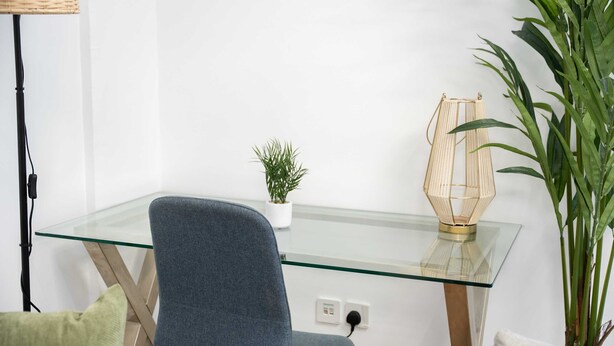 Een moderne kantoorruimte met een glazen tafel, een blauwe stoel, een kleine potplant, een vloerlamp en een grote groene plant.