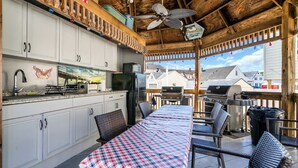 Une cuisine extérieure abritée, dotée d’un barbecue, d’une table à manger recouverte d’une nappe à carreaux, et offrant une vue sur les maisons au loin.