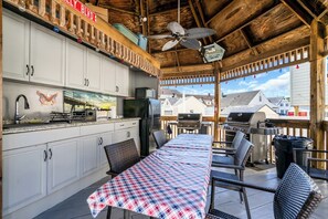 A covered outdoor kitchen with a grill, a dining table with a checkered tablecloth, and a view of houses in the background. - Professionally Managed | Walk to Beach (Seaside Heights)