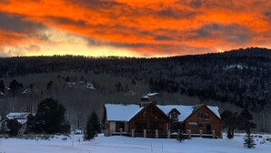 A log cabin with a snow-covered roof, surrounded by a snowy landscape and a forested hill. - Ski Chalet - Views, Hot Tub, Game Room (Angel Fire)