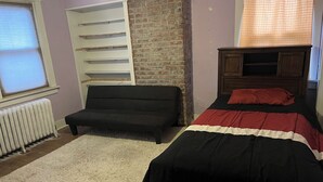 Ein Schlafzimmer mit einer Couch, einem Bett mit roter und schwarzer Bettdecke, einer Backsteinwand, einem Fenster mit Jalousien und einem Heizkörper.