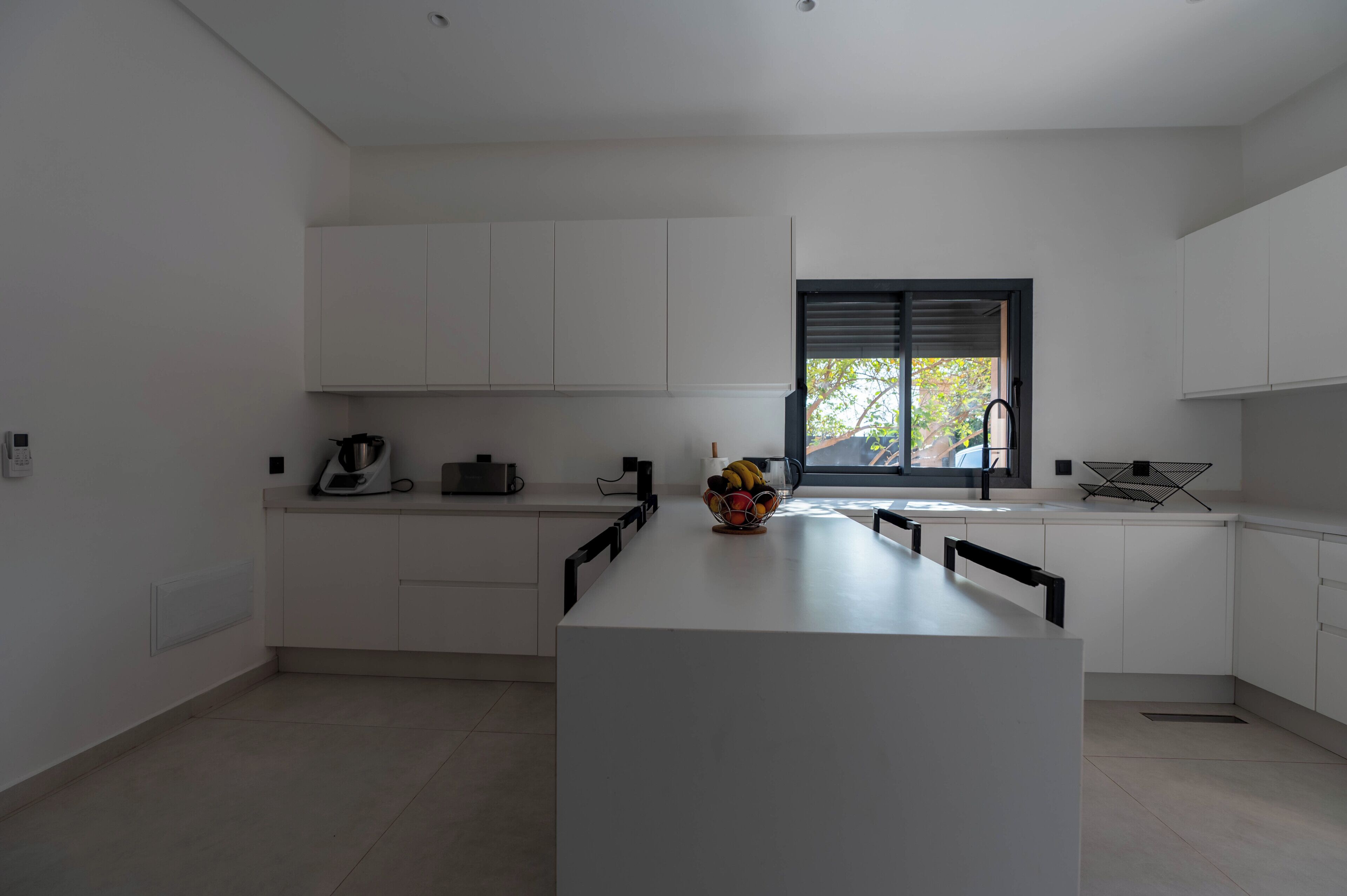A modern kitchen with white cabinetry, a central island, a window with blinds, and a fruit bowl on the island.