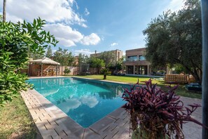 A swimming pool with a wooden deck, surrounded by greenery and a building in the background. - Villa Adam Luxe Marrakech, 5 Suites, Large Private Pool, sleeps 11 (Marrakech)
