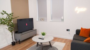Een moderne woonkamer met een flatscreen televisie, een ronde salontafel, een grijze bank met een oranje kussen, een kleine potplant en een wandlamp.
