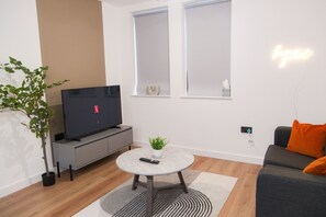 A modern living room with a flat-screen TV, a round coffee table, a grey sofa with an orange pillow, a small potted plant, and a wall-mounted light fixture. - Modern 1 bed flat in Birmingham city centre (Birmingham)