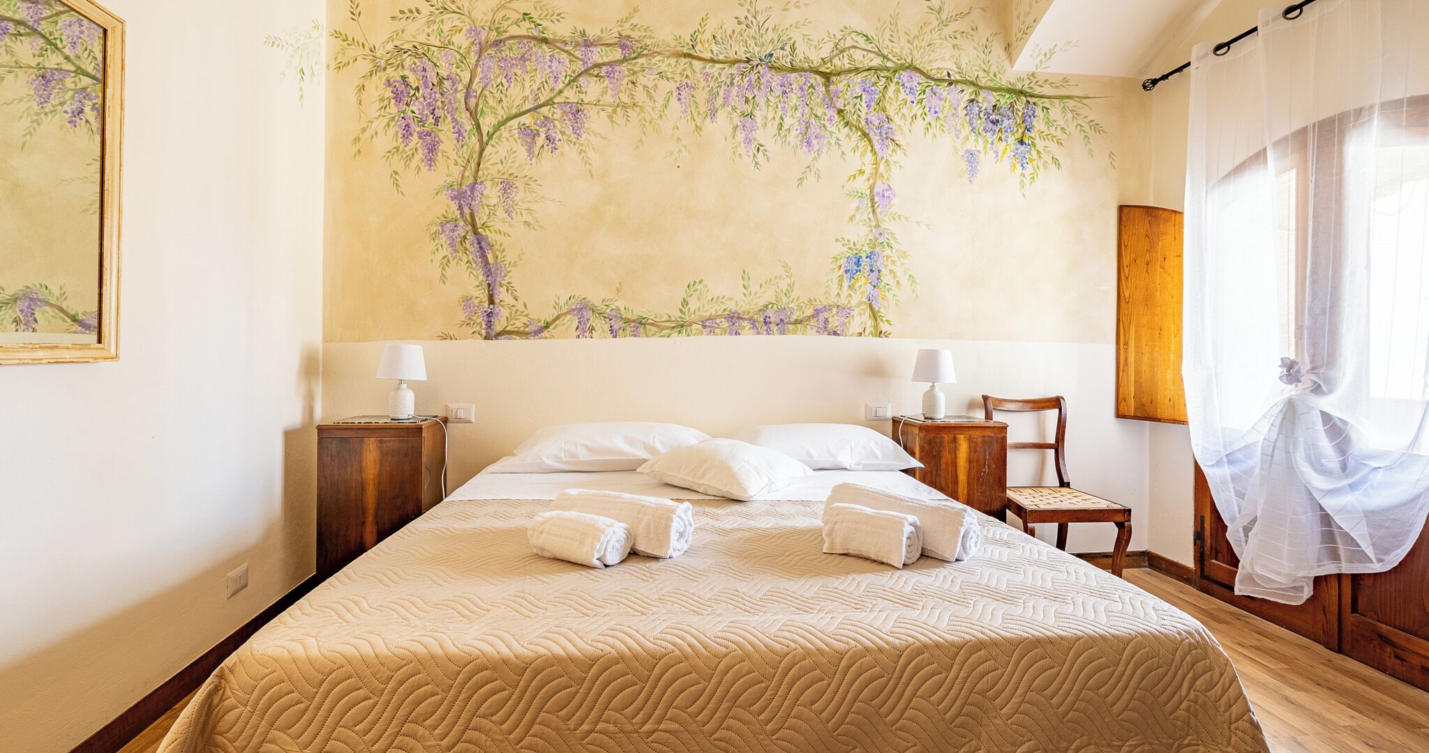 Une chambre d’hôtel avec un grand lit, deux tables de chevet, une chaise, une fenêtre avec des rideaux légers et une fresque murale.