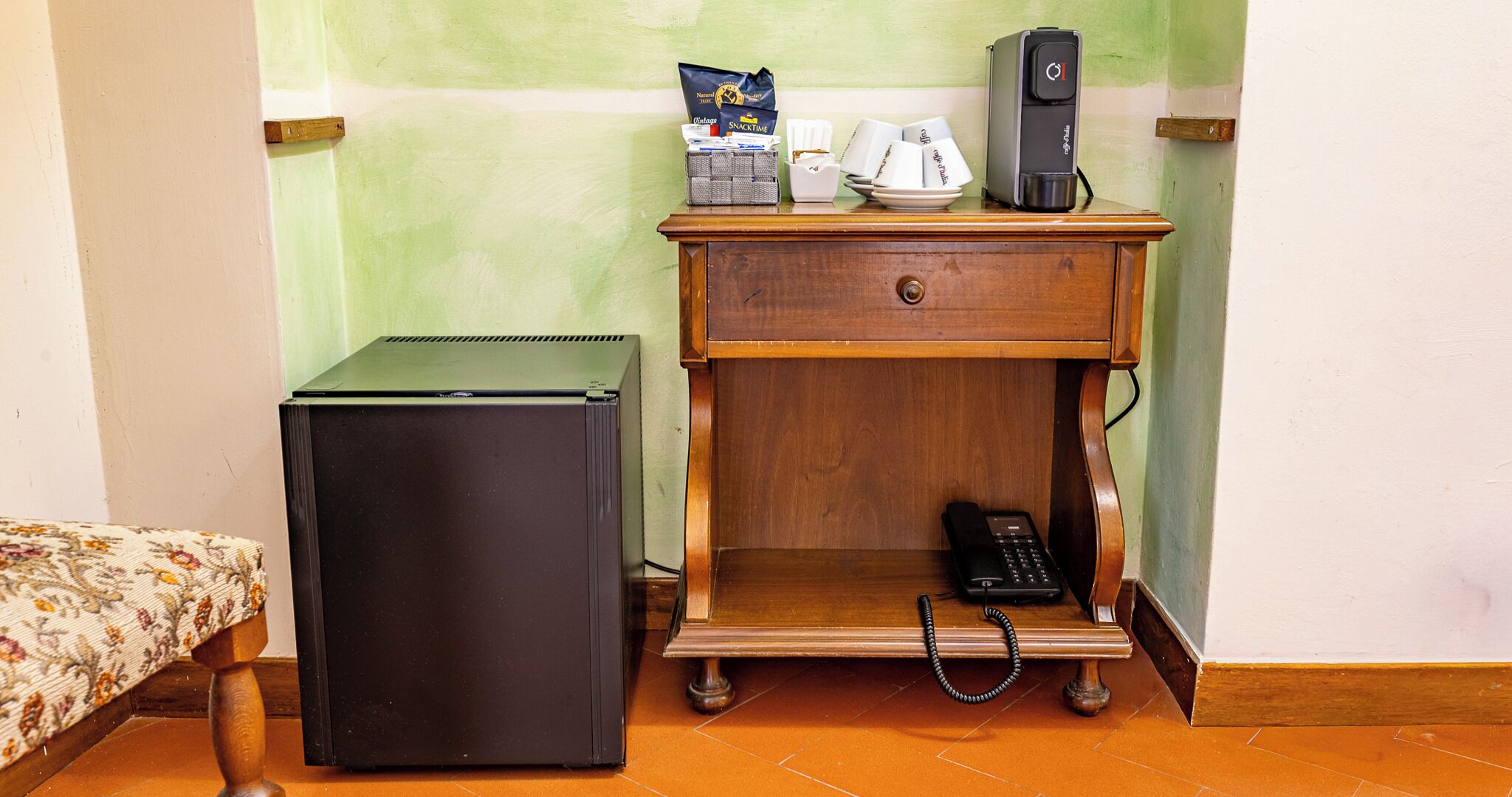 Une chambre de taille modeste, équipée d’un petit bureau en bois, d’un réfrigérateur, d’un téléphone et d’une machine à café.