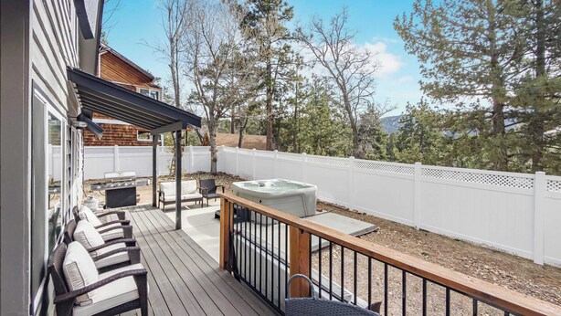 A deck with outdoor seating, a hot tub, and a view of trees and a white fence. - Bear Mesa Chalet | Hot Tub · Fireplace (Big Bear)