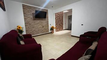 Ein Wohnzimmer mit Flachbildfernseher an einer strukturierten Wand, einer Sofagarnitur und einem Flur, der in einen weiteren Raum führt.