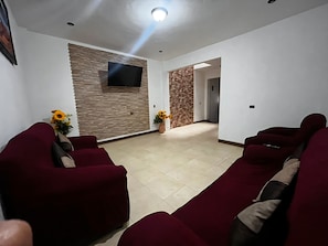 Een woonkamer met een flatscreen televisie aan een gestructureerde muur, een zithoek en een gang naar een andere kamer.