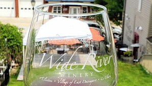 A wine glass with "Wide River Winery" text, set against a residential backdrop with a house, lawn, and distant buildings. - Charming Apartment in the heart of Downtown LeClaire sleeps 4 (Le Claire)