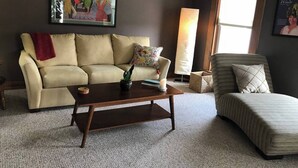 A living room with a beige sofa, a wooden coffee table, and a striped armchair. There is a painting on the wall and a tall floor lamp. - Charming Apartment in the heart of Downtown LeClaire sleeps 4 (Le Claire)