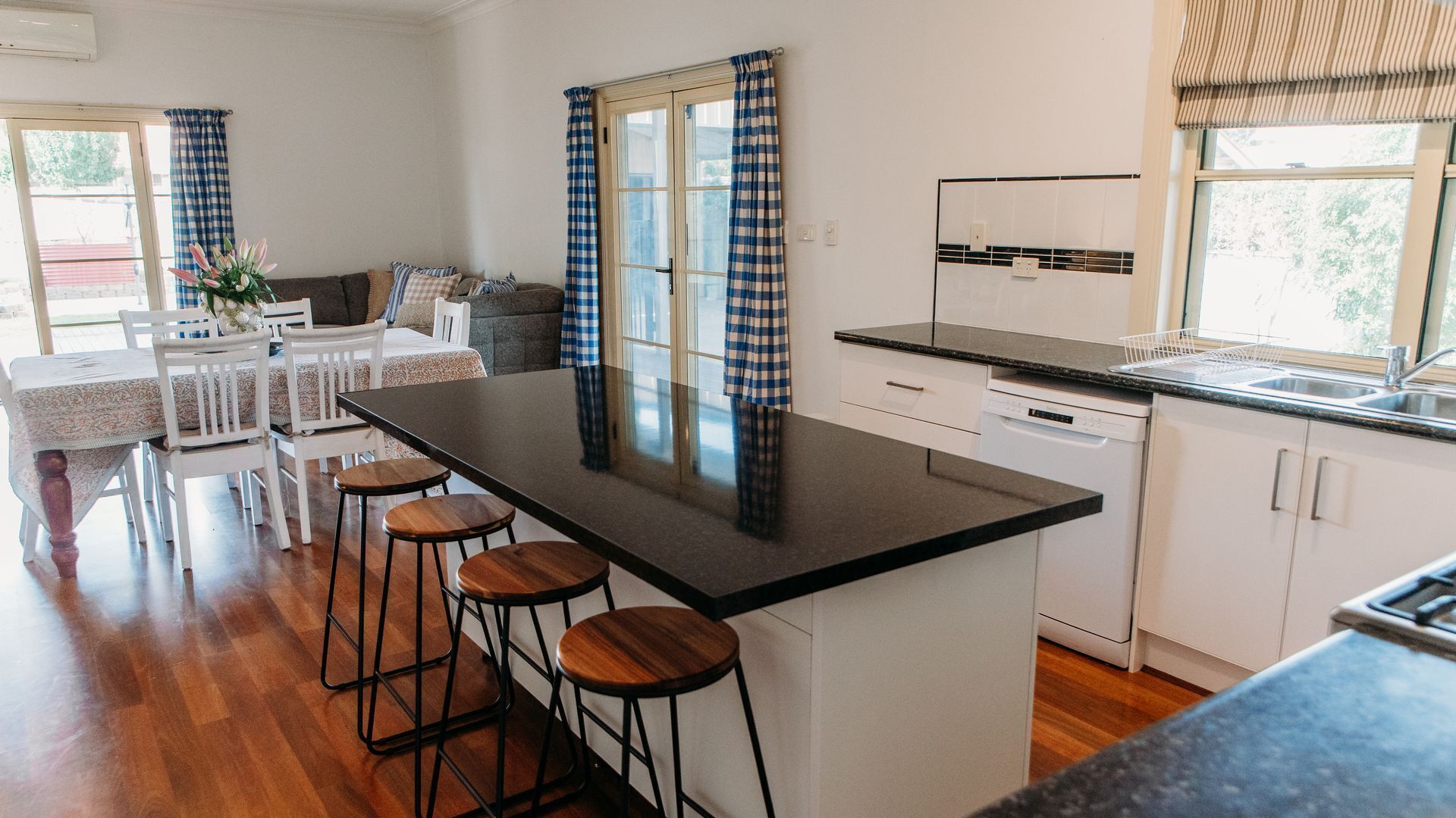 A modern kitchen with a central island, bar stools, a dining area with a table and chairs, and a view through the window.